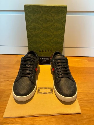 Gucci Ace sneakers  - Snygga Gucci Ace sneakers i svart med klassiskt GG-mönster, röda och blå ränder på sidan samt röd detalj på hälen. Skorna har vita sulor och svarta skosnören. Perfekta för dig som vill ha en lyxig och trendig look.
