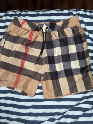 Burberry shorts - Snygga burrberry shorts med klassiskt rutigt mönster i beige, svart, vitt och rött. De har elastisk midja med svart dragsko och sidofickor. Perfekta för en trendig och avslappnad stil.