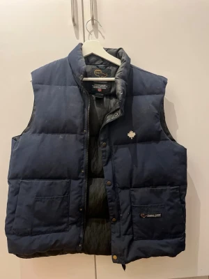 Mörkblå dunväst från Canada Goose - Snygg mörkblå dunväst från Canada Goose med klassisk passform och hög krage. Västen har två stora fickor framtill, tryckknappar och dragkedja. Ikonisk logga med lönnlöv på bröstet och märke på fickan. Perfekt för lager-på-lager-stil.