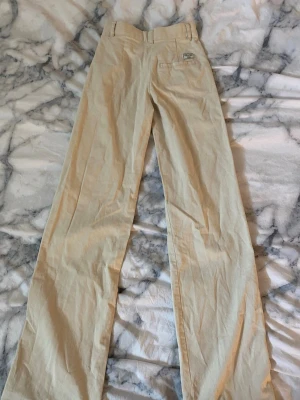 Beige raka kostymbyxor - Snygga beige kostymbyxor med raka ben och mellan hög midja. Byxorna har klassisk passform, bälteshällor och fickor fram. Materialet ser ut att vara bomull eller bomullsblandning, perfekt för en clean och stilren look.