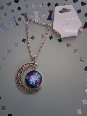 Halsband med måne och galaxmotiv - Snyggt halsband med en silverfärgad kedja och en hängande halvmåne i metall med detaljerat mönster. Inuti månen sitter en rund berlock med färgglatt galaxmotiv i blått, lila och vitt. Perfekt accessoar för dig som gillar rymd och unika smycken.
