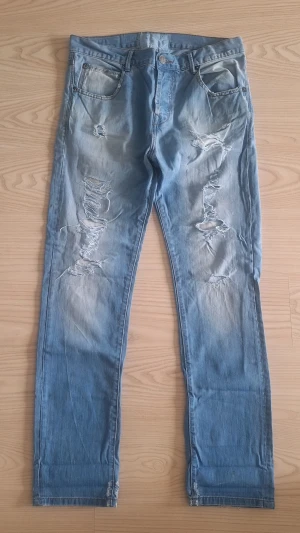 Ljusblå slitna jeans med hål stl W32 L32 - Säljer ett par ljusblå jeans med flera slitningar och hål på benen. Jeansen har klassisk femficksmodell och raka ben. Perfekta för en avslappnad och trendig streetstil. Materialet är denim i bomull.
