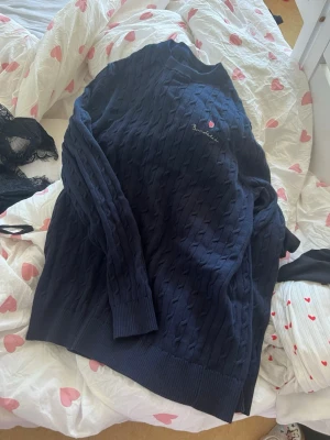 Mörkblå stickad tröja Bondelid - Mörkblå stickad tröja från Bondelid med diskret mönster och broderad logga på bröstet. Tröjan har rund halsringning, långa ärmar och ribbade muddar vid ärmslut och nederkant. Perfekt för en stilren och avslappnad look.
