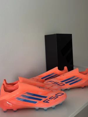 Adidas F50 CORAL BLACE Laceless Elite AG - Säljer ett par Adidas F50 fotbollsskor i en riktigt schysst orange färg med blå detaljer och logga. Skorna har en strömlinjeformad design utan snörning och är gjorda i syntetmaterial för bästa känsla och lätthet på planen. Perfekta för dig som vill sticka ut på matchen!