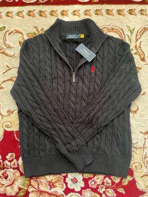 Grå kabelstickad tröja Polo Ralph Lauren - Kabelstickad grå tröja från Polo Ralph Lauren med halv dragkedja och hög krage. Tröjan har ett rött broderat logomärke på bröstet och ribbstickade muddar. Perfekt för en stilren och klassisk look.