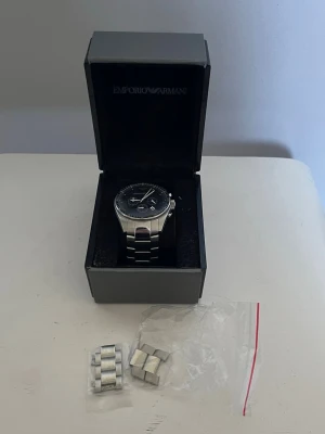 Emporio Armani silverfärgad klocka - Stilren klocka från Emporio Armani med svart urtavla och silverfärgat armband i metall. Klockan har kronograf-funktion och levereras i originalask med extra länkar. Perfekt accessoar för dig som gillar klassisk och modern design. Uret är ur funktion men går lätt och fixa på vilken klockaffär som helst (därav billigt pris)