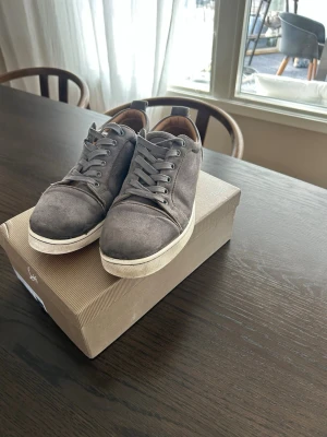 Gråa sneakers från Christian Louboutin - Snygga gråa sneakers från Christian Louboutin med klassisk låg profil och vita sulor. Skorna har ovandel i mocka och matchande gråa. Några små skador fram på skon men inget någon tänker på och fungerar fortfarande som de ska!