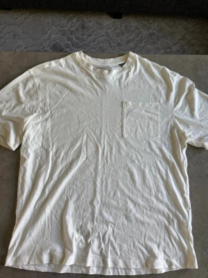 Vit t-shirt med bröstficka - En stilren vit t-shirt med rund halsringning och en praktisk bröstficka. Perfekt basplagg som passar till det mesta och är lätt att matcha. T-shirten har en klassisk passform och är tillverkad i mjuk bomull.