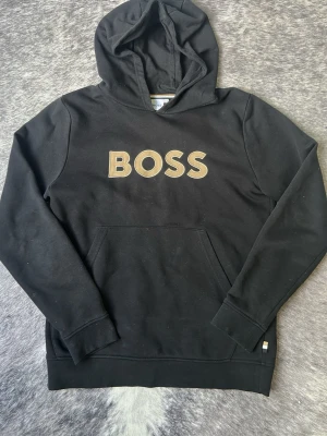 Svart hoodie från BOSS med logga - Svart hoodie från BOSS med stor guldfärgad logga på bröstet. Klassisk modell med huva och känguruficka framtill. Tillverkad i mjukt bomullsmaterial som är skönt att bära. Perfekt för en avslappnad och trendig stil.