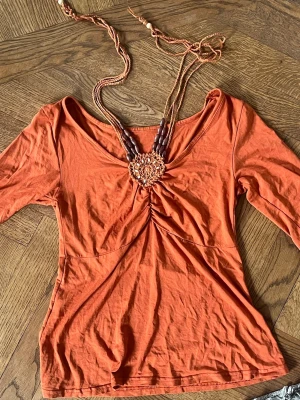Orange topp med smyckesdetalj - Orange topp med knytning i nacken och trekvarts ärm. Köpt second hand men aldrig använd av mig och inga defekter! Skulle säga storlek XS-S, skriv gärna vid frågor!!