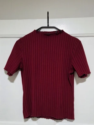 Ribbad vinröd topp från ARjen - Snygg vinröd ribbad topp från ARjen med korta ärmar och rak passform. Toppen har en enkel design med rund halsringning och är tillverkad i ett stretchigt material som sitter bekvämt. Perfekt att styla med jeans eller kjol.