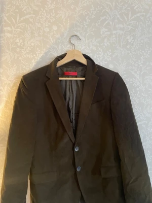 Mörkbrun kavaj från HUGO - Snygg mörkbrun kavaj från HUGO med klassisk passform och två knappar framtill. Kavajen har smala slag, två fickor och är helfodrad. Perfekt för en stilren look och passar till många olika outfits.