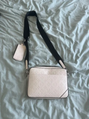 Vit axelväska från Louis Vuitton - Säljer en stilren vit axelväska från Louis Vuitton i präglat läder med klassiskt monogrammönster. Väskan har en bred svart axelrem och en matchande liten avtagbar miniväska. Perfekt för dig som vill ha en lyxig och trendig accessoar.