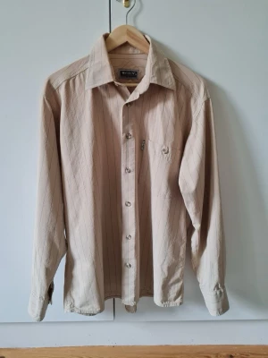 Beige randig skjorta från Mustang strl M - Stilren beige skjorta från Mustang med tunna vertikala ränder och klassisk krage. Skjortan har lång ärm, bröstficka och knäppning framtill. Finns ett litet omärkbart hål, se sista bilden. Storlek M.