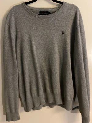 Grå tröja från Polo Ralph Lauren - Klassisk grå tröja från Polo Ralph Lauren med rund halsringning och diskret broderad logga på bröstet. Tröjan har långa ärmar och ribbade muddar vid ärmslut och nederkant. Perfekt för en stilren och enkel look. Perfekt basplagg där man kan lägga på fler lager för att få en bra look