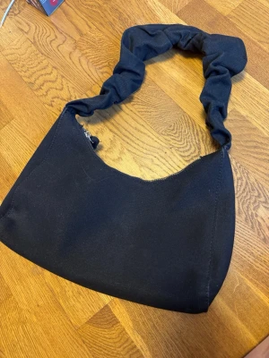 Svart axelväska med scrunchie-band - Trendig svart handväska i tyg med scrunchie-inspirerat axelband och dragkedja upptill. Väskan har en enkel, stilren design och ett innerfack med dragkedja för småsaker. Perfekt för dig som gillar en clean look och vill ha plats för det viktigaste.
