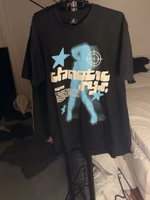 Svart t-shirt med blått tryck - Cool svart t-shirt med stort blått och vitt tryck framtill. Motivet har en siluett, stjärnor och texten 'chaotic ryr'. T-shirten har rund hals och korta ärmar, perfekt för en streetwear-look.