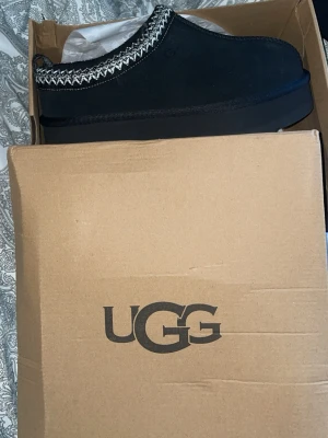 Svarta UGG Tazz tofflor - Säljer ett par svarta UGG Tazz tofflor i mocka med tjock sula och dekorativt mönster i vitt och grått runt kanten. Tofflorna har en modern chunky design och är fodrade för extra komfort. Perfekta för att hålla fötterna varma och stilrena inomhus.