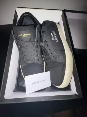 Saint Laurent svarta sneakers - Svarta sneakers från Saint Laurent med vit sula och handskriven logga på sidan. Skorna har klassisk låg profil, svarta skosnören och ovandel i canvas. Perfekta för dig som gillar stilrena och exklusiva detaljer.