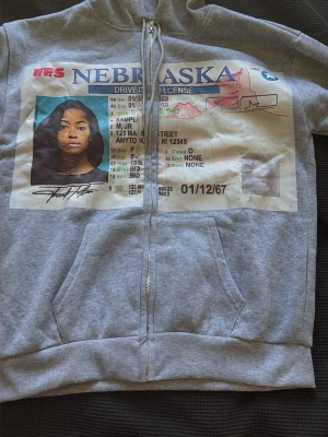 Grå hoodie med Nebraska-tryck - Grå zip-hoodie med stor, färgglad print av ett Nebraska körkort på framsidan. Hoodien har huva med dragsko och framfickor. Materialet är mjukt och bekvämt, perfekt för en avslappnad streetwear-look.