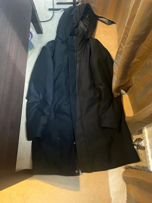 Svart regnjacka från Uniqlo med huva - Snygg svart regnjacka från Uniqlo i storlek XS. Jackan har en minimalistisk design med huva och dragkedja framtill, samt tryckknappar. Tillverkad i vattenavvisande material som passar perfekt för regniga dagar.