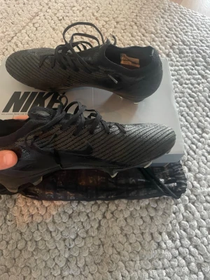 Nike zoom vapor 15 elite fg fotbollsskor  - Snygga svarta Nike Zoom vapor 15 fotbollsskor med stickad ovandel och klassisk snörning. Skorna har en strumpliknande passform och mönstrad yta för bättre bollkontroll. Perfekta för dig som vill ha stilrena och snabba skor på planen.