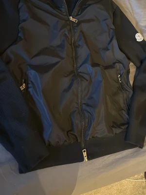 Svart bomberjacka från Moncler - Snygg svart bomberjacka från Moncler med ribbade ärmar och muddar. Jackan har två fickor med dragkedja och en klassisk Moncler-logga på ena ärmen. Materialet är glansigt och ger en modern look. Perfekt för dig som vill ha en stilren och trendig jacka. OBS! Har några skavanker som visas på de två sista bilderna!