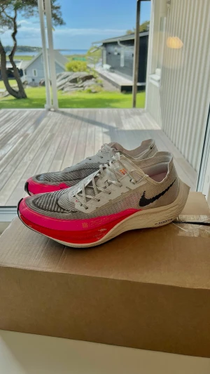 Nike ZoomX Vaporfly Next% sneakers - Nike ZoomX Vaporfly Next% sneakers i grått och neonrosa med svart swoosh. Ovandel i mesh för extra ventilation och lätt känsla. Mellansula med ZoomX-dämpning för maxad energiåtergivning. Perfekta för dig som vill ha snabba och snygga sneakers.