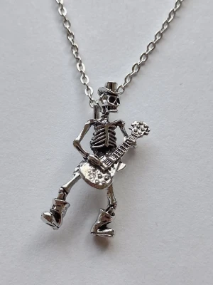 Halsband med skelett och gitarr - halsband i silverfärgad rostfritt metall med ett coolt skelett som spelar elgitarr och har hatt och boots. Kedjan är tunn ankarlänk. Perfekt accessoar för dig som gillar rockig stil och detaljerade smycken.