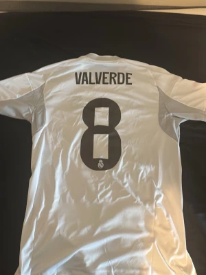 Real Madrid matchtröja Valverde 8 -  vit Real Madrid matchtröja från Adidas med Valverde och nummer 8 på ryggen. Tröjan har klubbmärke, sponsortryck och svarta detaljer på axlarna samt gula linjer längs sidorna. Helt ny från unisports hemsida