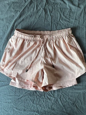 Ljusrosa träningsshorts med snörning - Snygga ljusrosa/ laxrosa träningsshorts med elastisk midja och snörning framtill. Shortsen har en lös passform och är tillverkade i ett lätt syntetmaterial som andas. Har underbyxor som sitter lite tightare på benen, men med en elastisk och mjuk känsla. Perfekta för aktiva dagar eller avslappnade stunder.