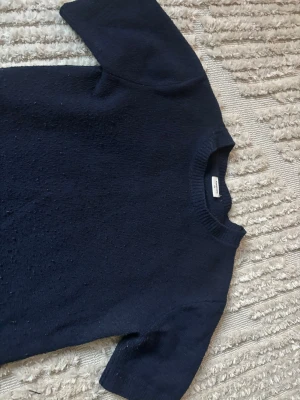 Mörkblå stickad tröja Kappahl - Mörkblå stickad tröja från Kappahl med korta ärmar och rund halsringning. Tröjan har en enkel och stilren design med ribbstickad kant vid halsen. Perfekt för lager-på-lager och passar till många olika outfits.