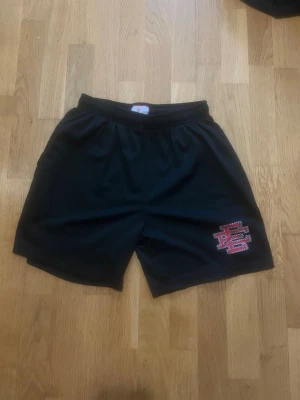 Svarta meshshorts med röd logga - Svarta shorts i meshmaterial med elastisk midja och en röd-vit broderad logga på vänster ben. Perfekta för sport eller chill, med luftig känsla och klassisk passform.