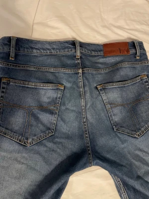 Blå jeans med raka ben - Säljer ett par klassiska blå jeans från tiger of sweden med raka ben och fem fickor. Jeansen har en snygg tvättad look, kontrastsömmar och en brun läderpatch bak i midjan. Perfekta till vardagsstilen och passar till det mesta. Materialet är denim i bomull.