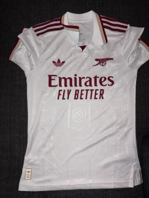 Arsenal vit matchtröja Adidas - Säljer en vit Arsenal matchtröja från Adidas med röda detaljer och klubbmärke på bröstet. Tröjan har trycket 'Emirates Fly Better' och klassiska Adidas-ränder på axlarna. Materialet är lätt och andas, perfekt för fotboll eller träning.