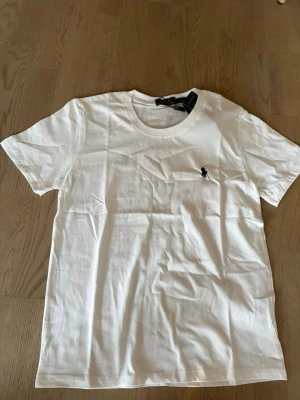 Vit t-shirt från Polo Ralph Lauren - Klassisk vit t-shirt från Polo Ralph Lauren med rund halsringning och ikonisk broderad logga på bröstet. Tillverkad i mjuk bomull och har en normal passform. Perfekt basplagg för en stilren och enkel look.