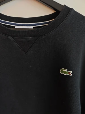 Svart sweatshirt från Lacoste XL - Klassisk svart sweatshirt från Lacoste med rund halsringning och den ikoniska gröna krokodilloggan broderad på bröstet. Tröjan har ribbade muddar vid ärmslut och nederkant samt en snygg detalj vid halsen. Perfekt för en avslappnad och stilren look.