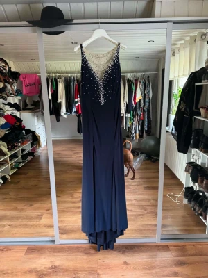 Marinblå långklänning med pärlor - Elegant marinblå långklänning med djup v-ringning och meshdetalj framtill. Klänningen är dekorerad med vita pärlor som ger en lyxig känsla. Perfekt för speciella tillfällen där du vill sticka ut.