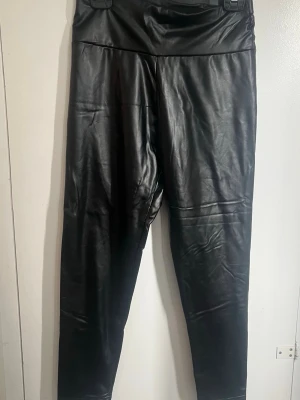 Svarta leggings i fuskskinn - Snygga svarta leggings i glansigt fuskskinnmaterial med hög midja. Tight passform och stretchigt tyg som ger en cool look. Perfekta att styla med oversized tröja eller crop top.