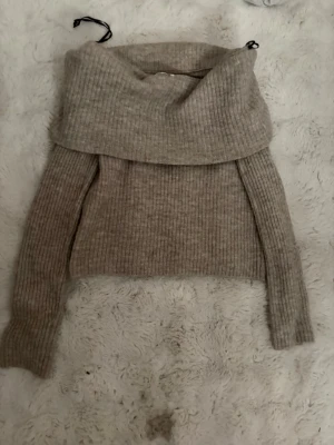 Beige ribbstickad off shoulder-tröja - Säljer en beige ribbstickad tröja med bred off shoulder-krage. Tröjan har långa ärmar och är gjord i ett mjukt, tjockt material som passar perfekt för kyligare dagar. Enkel att styla med jeans eller kjol för en trendig look.