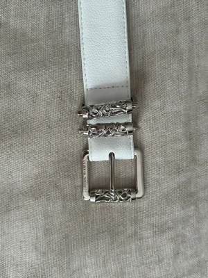 Vitt roller belt - Stilrent vitt skärp i skinn med präglade korsdetaljer längs remmen och snyggt dekorerat silverspänne. Perfekt accessoar för att lyfta din outfit med en cool touch. Bältet har bred design och detaljerade metalldelar. 110 cm