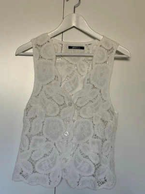 Vit virkad väst från Gina Tricot - Superfin vit väst från Gina Tricot med v-ringad hals och knappar framtill. Västen har ett broderat blommönster och är ärmlös med vågig nederkant. Perfekt att styla över en enkel topp för en trendig look.