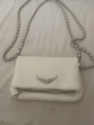 Vit handväska med kedjerem - Snygg vit handväska med silverfärgad kedjerem och dekorativt emblem med vingar framtill. Väskan har en dragkedja på framsidan och är tillverkad i ett slätt material. Perfekt för att lyfta din outfit med en cool detalj.
