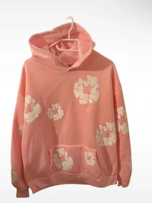 Denim tears - Säljer en oversized rosa hoodie med vita blommönster över hela tröjan. Hoodien har stor huva och en klassisk magficka framtill. Perfekt för dig som gillar färgglada och unika plagg. Materialet är mjukt och skönt, troligtvis bomullsblandning.