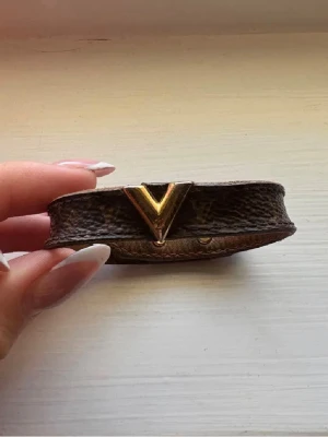 Louis Vuitton brunt läderarmband - Perfekt accessoar för att lyfta din outfit.Snyggt brunt armband från Louis Vuitton i äkta läder med klassiskt LV-mönster och guldfärgad V-detalj framtill. Armbandet har justerbar knäppning med två guldfärgade nitar och är perfekt för att ge din outfit en lyxig touch. Den är väl använd ! Och har lite små defekter där man stänger arm