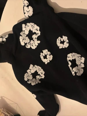 Svart denim tears Hoodie - Cool svart hoodie med vita blommönster tryckta över hela tröjan, inklusive huvan och ärmarna. Hoodien har en klassisk känguruficka framtill och en stor huva. Perfekt för dig som vill sticka ut med en unik och trendig stil.