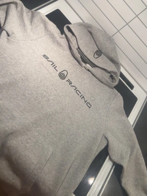 Grå hoodie från Sail Racing - Snygg grå hoodie från Sail Racing med svart logga tryckt på bröstet och en extra logga på huvan. Klassisk känguruficka framtill och mjukt material som känns skönt mot huden. Perfekt för dig som gillar stilren och sportig design.