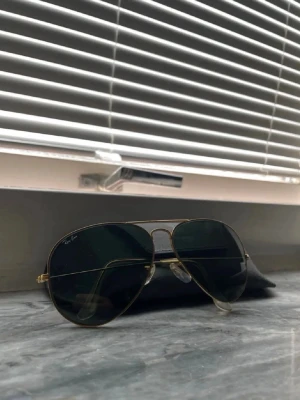 Ray-Ban aviator solglasögon guld - Klassiska Ray-Ban aviator solglasögon med tunna guldfärgade metallbågar och mörkgröna linser. Snygg och tidlös modell som passar till det mesta. Kommer med ett svart fodral. 