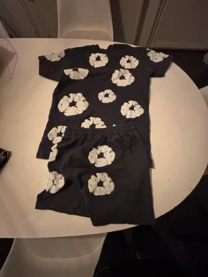 Svart set denim tears - Säljer ett svart tvådelat set med t-shirt och shorts, dekorerat med stora vita blommönster över hela plaggen. Setet har en avslappnad passform och elastisk midja på shortsen. Perfekt för dig som gillar unika och lekfulla mönster.