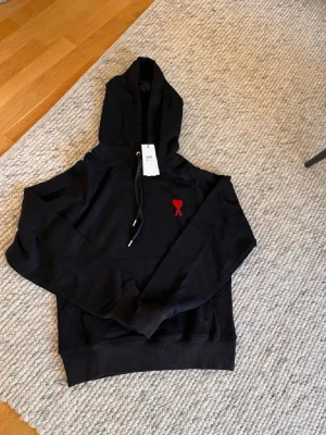 Svart hoodie från Ami Paris - Svart hoodie från Ami Paris med röd broderad logga på bröstet. Klassisk modell med huva och dragsko samt magficka framtill. Tillverkad i mjukt material som passar perfekt för en avslappnad stil.